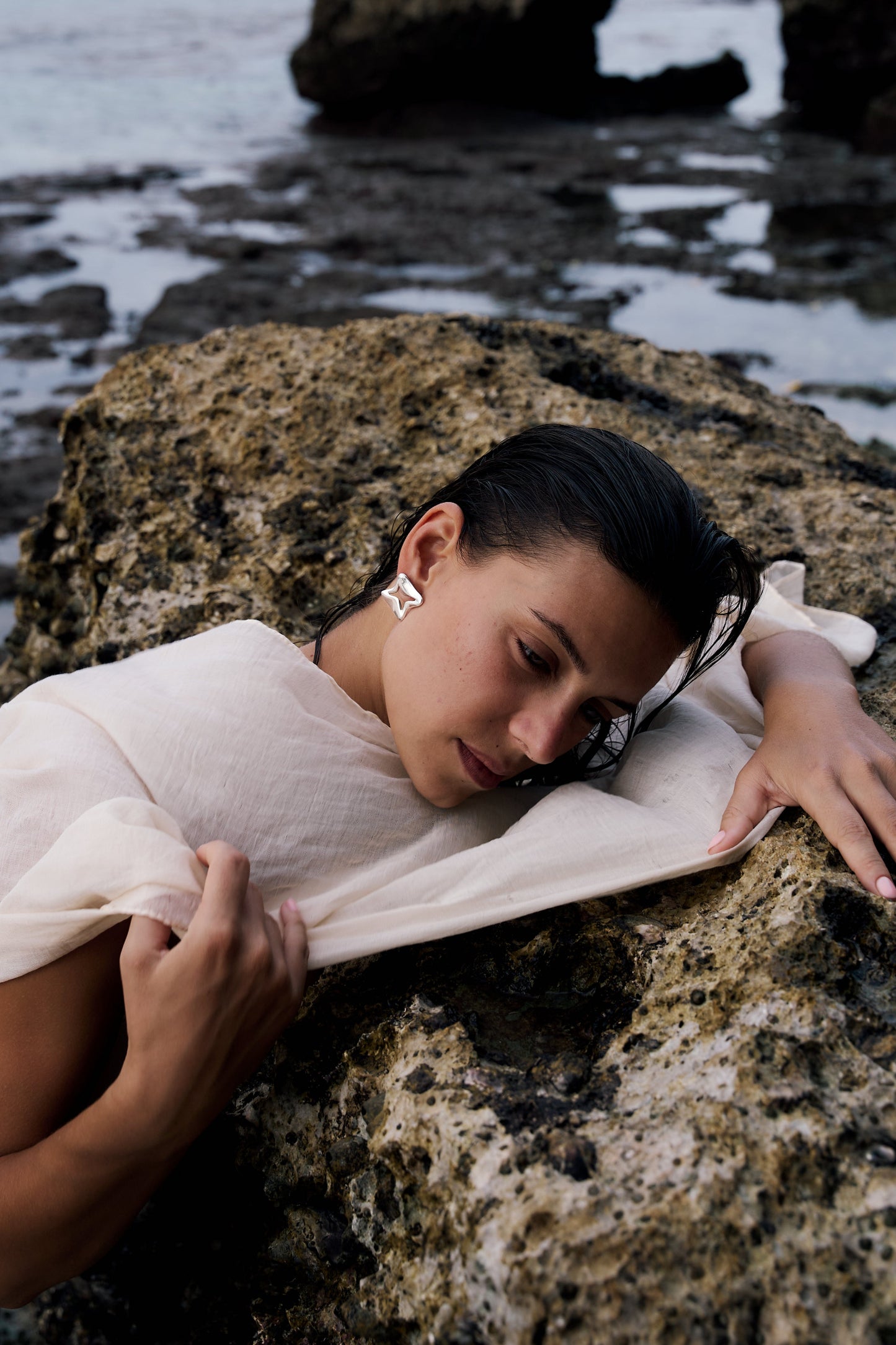 Woman lying on a rock by the water, wearing a light-colored dress.